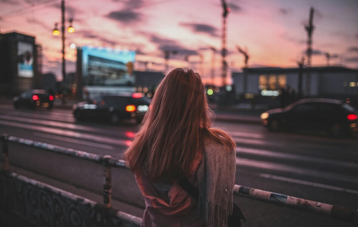 A solo traveler stands by the roadside at sunset, gazing at the bustling city traffic and illuminated billboards