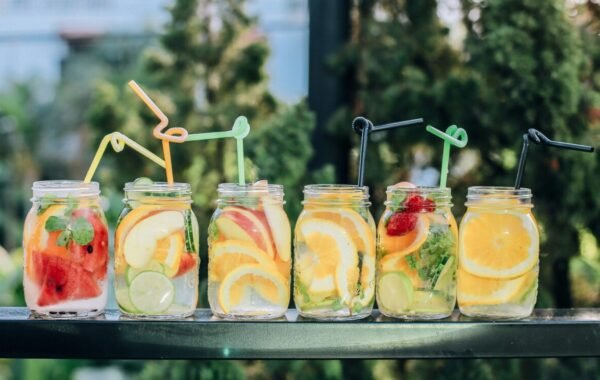 Colorful summer fruit-infused drinks in jars, featuring vibrant slices of watermelon, apple, orange, and lime, garnished with mint leaves, displayed in a row with colorful straws against a lush green background