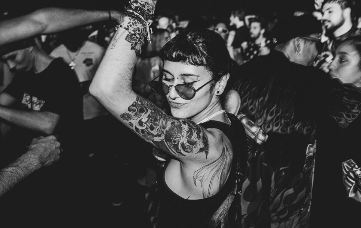 A stylish individual with tattoos dances in a lively crowd, wearing sunglasses and showcasing intricate body art on their arm, capturing the vibrant energy of a nightlife scene