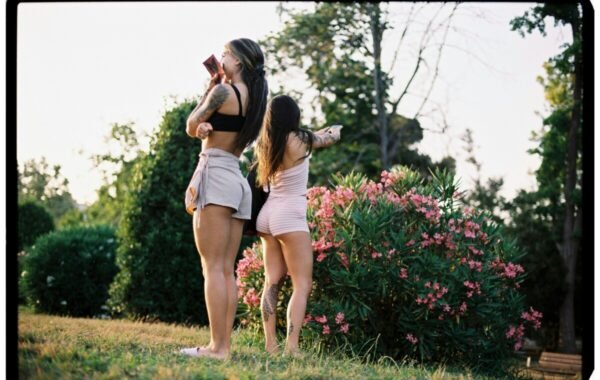 Two women showcasing their tattoos while enjoying a sunny day in a garden, surrounded by blooming flowers and greenery