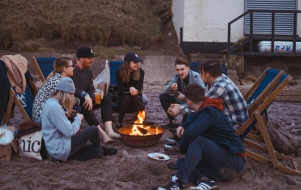 Group of friends gathered around a campfire on the beach, sharing stories and enjoying drinks, with lounge chairs set up nearby