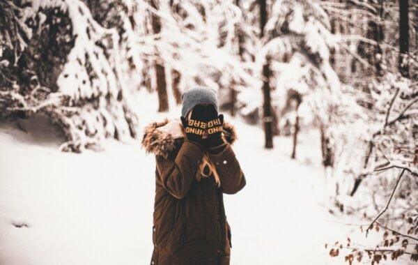 A solo traveler wearing a winter coat and beanie stands in a snowy forest, playfully covering their face with gloves, capturing the essence of adventure in nature