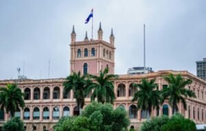 Historic government building in Paraguay featuring the national flag and surrounded by lush palm trees against a cloudy sky