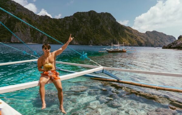Young man enjoying a coconut drink while sitting on a boat in clear turquoise waters, surrounded by lush mountains in the Philippines