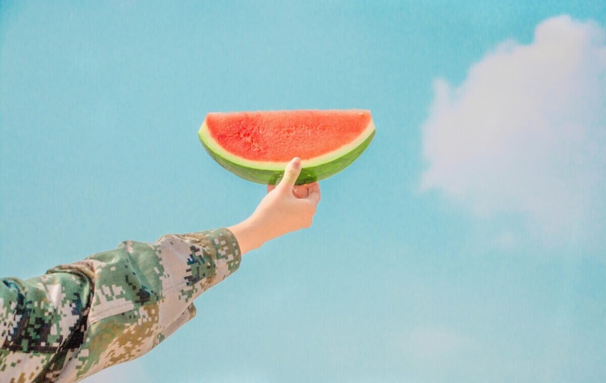 A hand in a camouflage sleeve holds a slice of watermelon against a bright blue sky with fluffy clouds, capturing the essence of summer