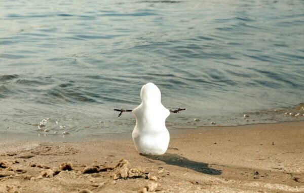 A small snowman stands on the beach, facing the water, with arms made of twigs, illustrating a whimsical contrast between winter and summer