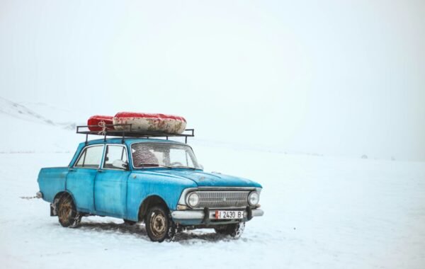 A vintage blue car parked on a snowy landscape with a red kayak on the roof under a cloudy sky