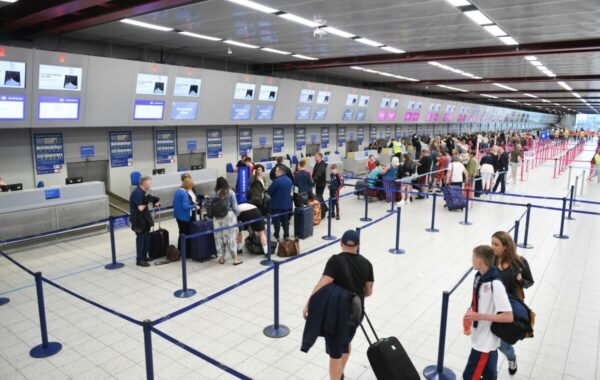 Crowded airport check-in area with passengers in line, multiple airline counters visible, and digital displays overhead indicating flight information