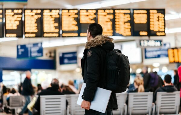 Person with a backpack looking at flight information boards in a busy airport terminal, with travelers seated in the background