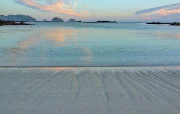 Serene beach scene with smooth sandy shore, calm turquoise waters, and distant rocky islands under a pastel sky