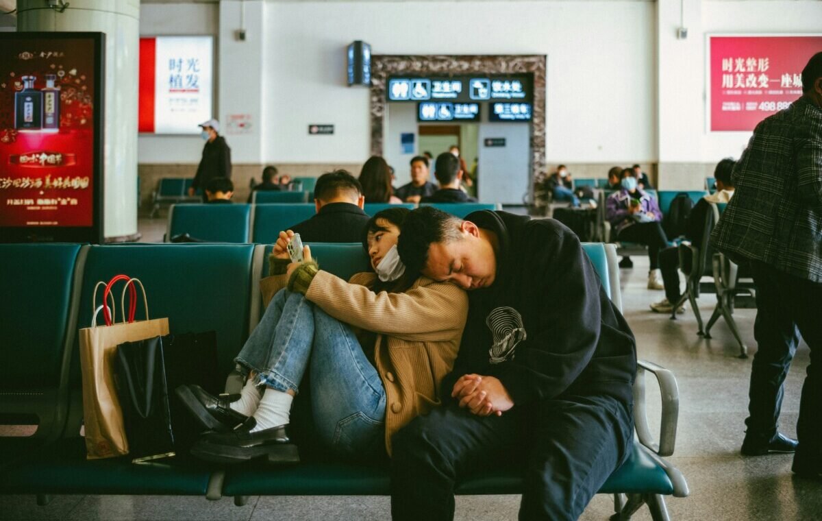 A couple rests together on a bench in a busy airport terminal, surrounded by other travelers