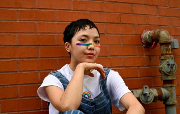 Young person with short dark hair, wearing overalls and a white t-shirt, poses against a brick wall