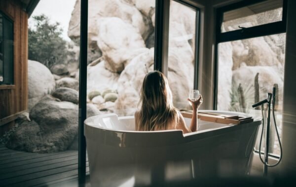 A woman relaxes in a luxury freestanding bathtub indoors, holding a glass while enjoying a serene view of boulders and greenery through large windows