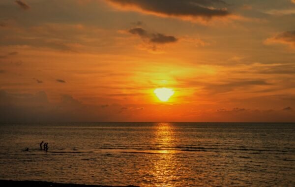 Scenic sunset view over the ocean in Bali, featuring silhouetted figures wading in the water as the sun sets on the horizon, casting golden reflections across the sea