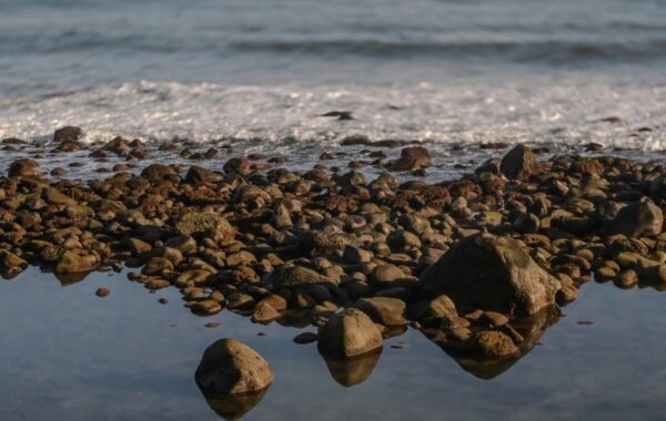 Wet rocky beach at sunset in Bali, featuring smooth stones partially submerged in shallow water, with gentle waves lapping at the shore