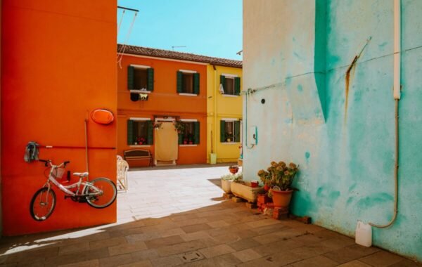 Colorful alleyway in Burano, Italy, featuring vibrant orange and turquoise walls, a bicycle hanging on the wall, and charming flower pots, showcasing the island's unique aesthetic