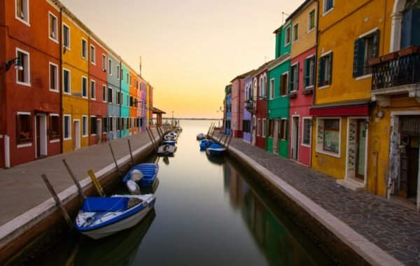 Colorful houses line the canals of Burano Island in Italy at sunset, with boats moored along the tranquil waterway, showcasing the island's vibrant charm