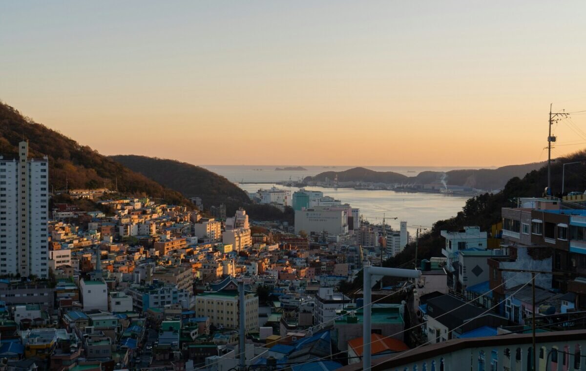 A stunning sunset view of Busan, South Korea, showcasing colorful buildings on the hillside overlooking the harbor and mountains in the distance
