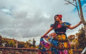 Woman wearing a colorful traditional Colombian dress, adorned with intricate floral patterns, poses against a dramatic cloudy sky in a picturesque outdoor setting