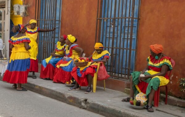 Vibrant scene of women dressed in colorful traditional Colombian attire, engaging in conversation and selling fruits along a street in a lively neighborhood