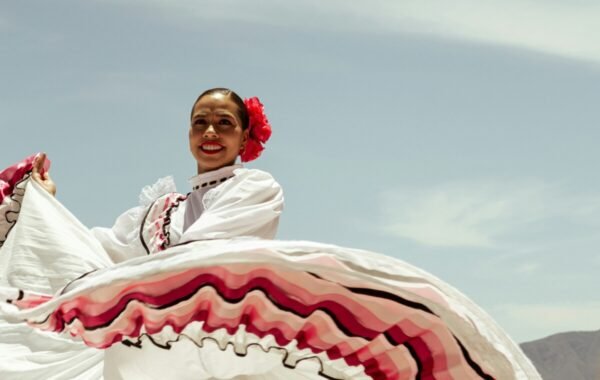 A woman in traditional Colombian attire joyfully dancing with a flowing white dress adorned with pink and black stripes, set against a clear blue sky