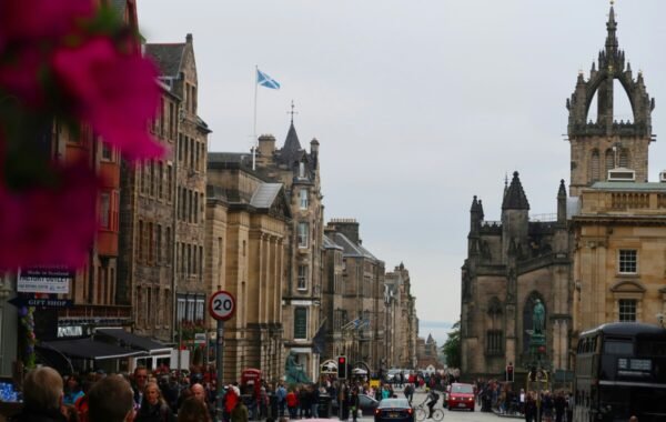 Bustling street scene in Edinburgh showcasing historic architecture, a Scottish flag, and crowds of pedestrians, along with notable buildings and traffic