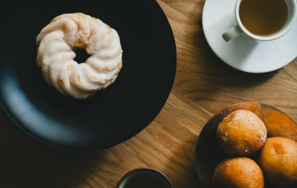 A top view of a glazed donut on a black plate, accompanied by golden pastry balls on a wooden table, with a cup of tea nearby