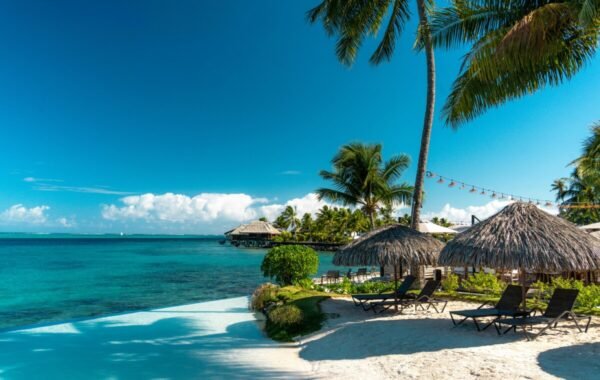 Tropical beach in French Polynesia featuring thatched umbrellas, palm trees, and a serene turquoise lagoon under a clear blue sky