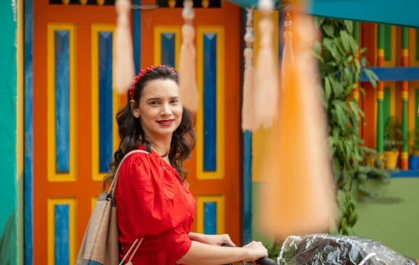 A woman in a red dress smiles in front of vibrant orange doors in Guatapé, Colombia, surrounded by colorful decor and hanging tassels