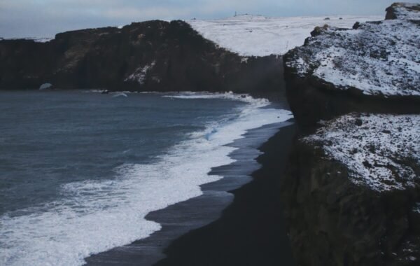 Snow-covered cliffs meet a dark beach as ocean waves crash along the shore in Iceland, showcasing the dramatic coastal landscape and winter scenery