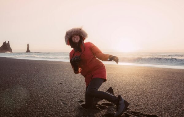 A person in a red coat and fur-lined hood enjoys a beach in Iceland, kneeling on black sand with a camera in hand, as the sun sets in the background