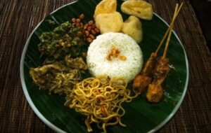 A vibrant plate of traditional Indonesian cuisine featuring jasmine rice, spicy chicken satay skewers, sautéed vegetables, fried noodles, peanuts, and crispy snacks, all presented on a banana leaf
