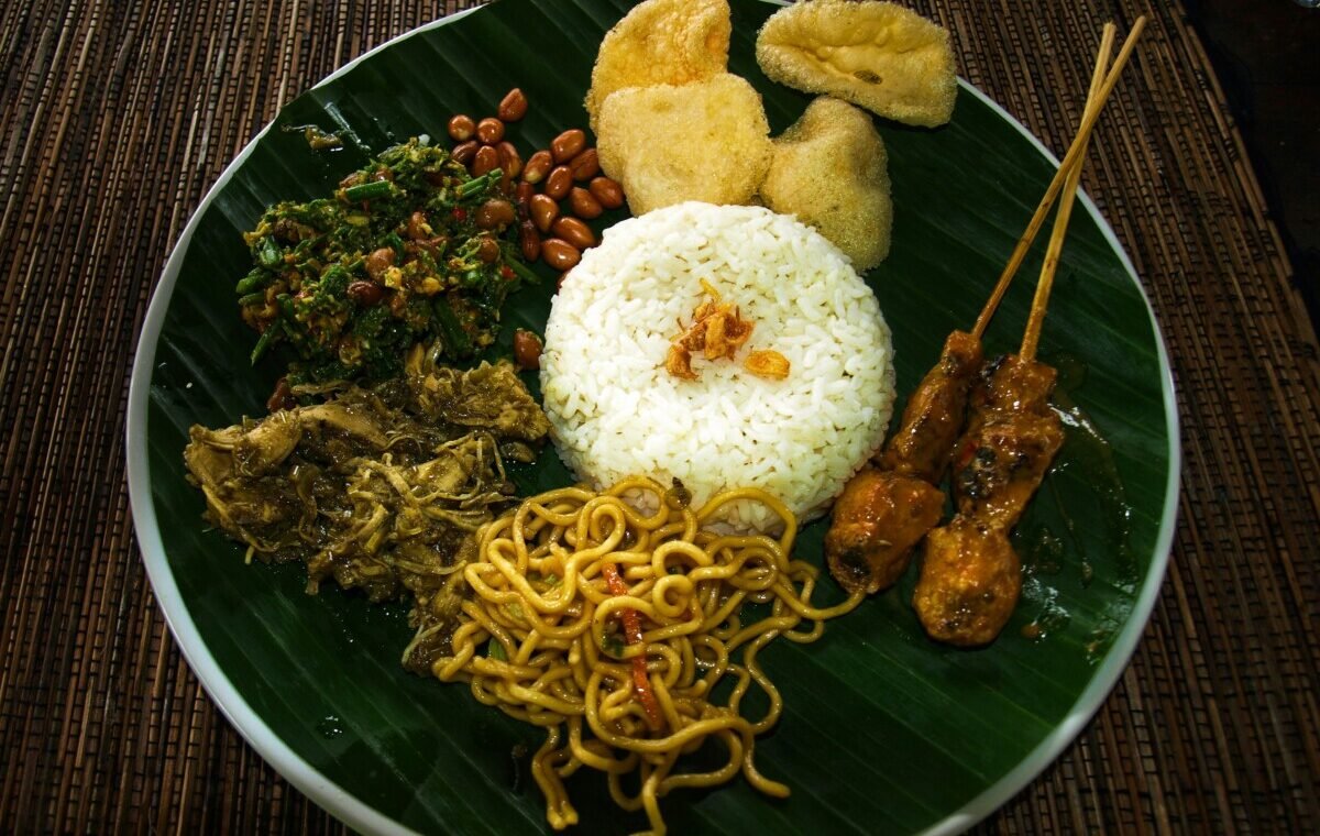 Traditional Indonesian cuisine featuring a vibrant platter with white rice, satay skewers, fried noodles, and assorted vegetables, served on a banana leaf