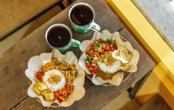 Two bowls of traditional Indonesian dishes featuring rice, assorted meats, and fresh vegetables, accompanied by two cups of dark coffee, set on a wooden table