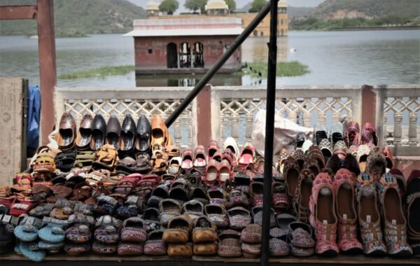 Colorful traditional footwear is displayed on a market stall by the serene waters of Jaipur, showcasing a variety of handcrafted shoes set against a picturesque backdrop of a historical structure and rolling hills