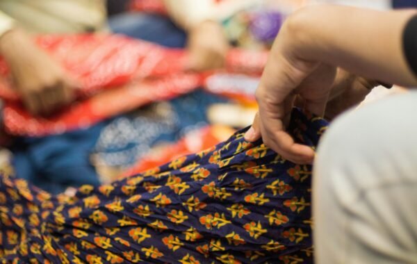 A close-up of a hand holding the textured fabric of a traditional Jaipur garment, surrounded by brightly colored textiles in a lively market setting