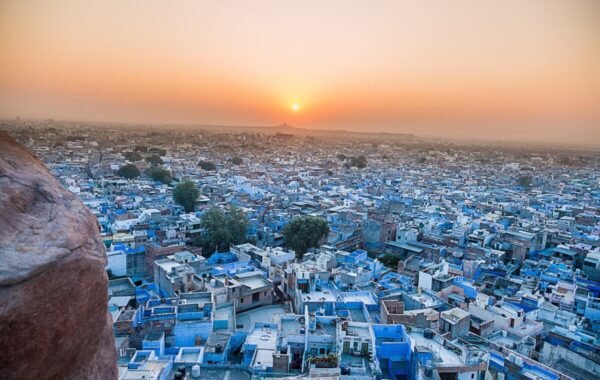 Aerial view of Jodhpur, India, showcasing the city's iconic blue buildings at sunset, with a warm orange glow illuminating the horizon