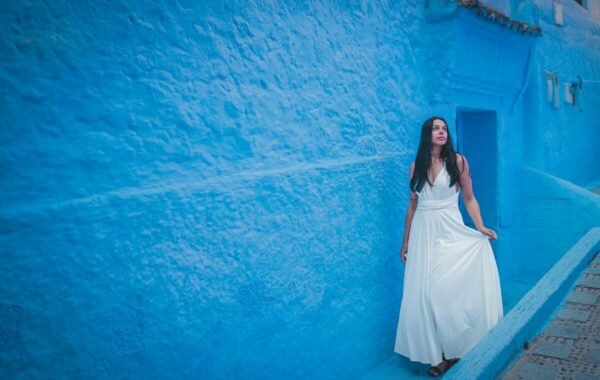 A woman in a flowing white dress stands gracefully against the vibrant blue walls of Morocco's Chefchaouen, highlighting the city's famous blue hue