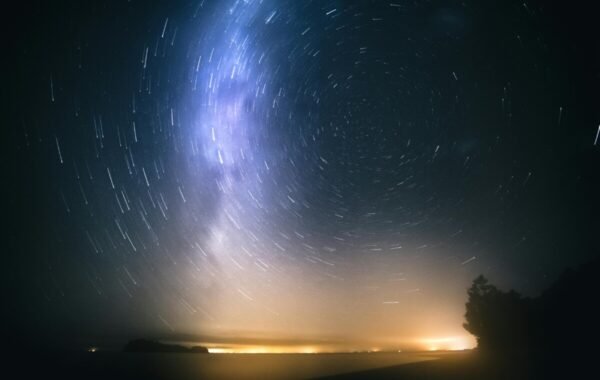 Starry night sky over New Zealand, showcasing the Milky Way and star trails above a tranquil beach with distant lights on the horizon