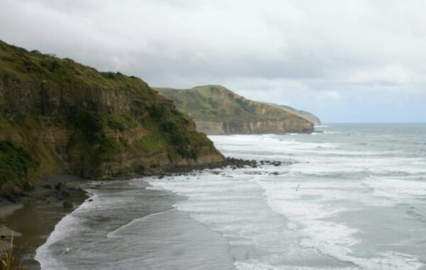 Scenic view of New Zealand coastline featuring rugged cliffs, crashing waves, and a cloudy sky, showcasing the natural beauty of the landscape