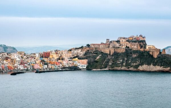 View of Procida Island, Italy, showcasing colorful buildings along the shoreline with a castle perched on a rocky hill in the background, under a cloudy sky