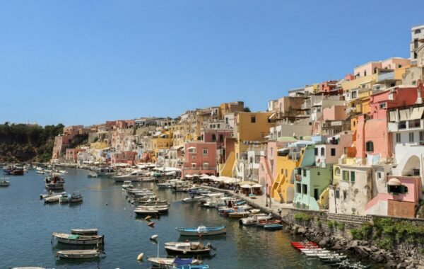Colorful buildings line the coastal harbor of Procida Island, Italy, with boats floating in the serene waters under a clear blue sky