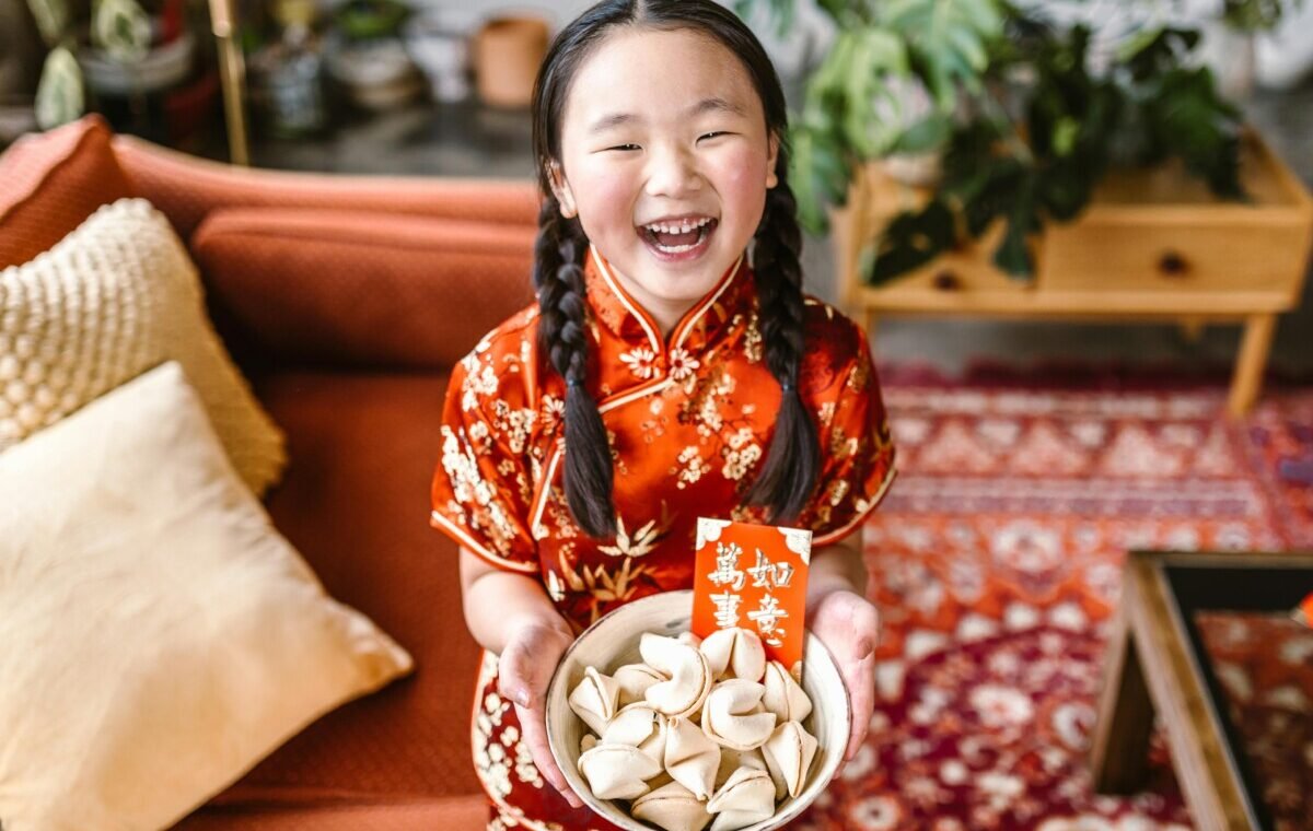 A joyful child in a traditional red Chinese outfit holds a bowl of dumplings, symbolizing prosperity, while beaming with happiness in a cozy living room setting