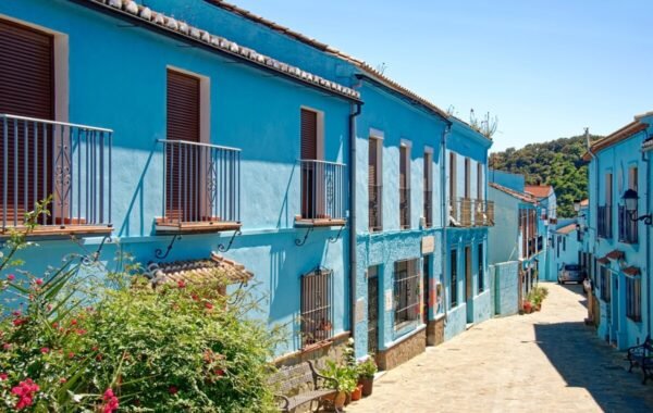 Picturesque blue buildings line a charming cobblestone street in Spain, adorned with vibrant flowers and greenery under a clear sky
