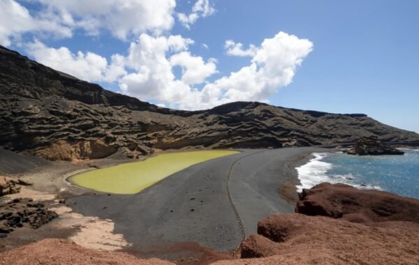 A breathtaking view of a volcanic landscape in Spain, featuring a striking green lagoon surrounded by dramatic black volcanic rock formations and a clear blue sky