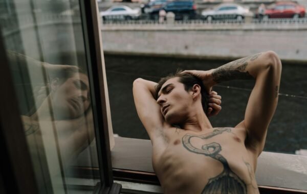 Young man with tattoos relaxing by a window, reflecting in glass, with a view of a river and parked cars in the background
