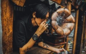 Individual with tattoos enjoying a cigarette while leaning out of a vintage shop window, surrounded by retro decor including a gramophone