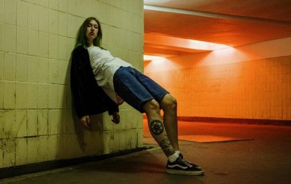 Person with green hair leaning against a wall in an underground setting, wearing a black jacket, white t-shirt, and denim shorts