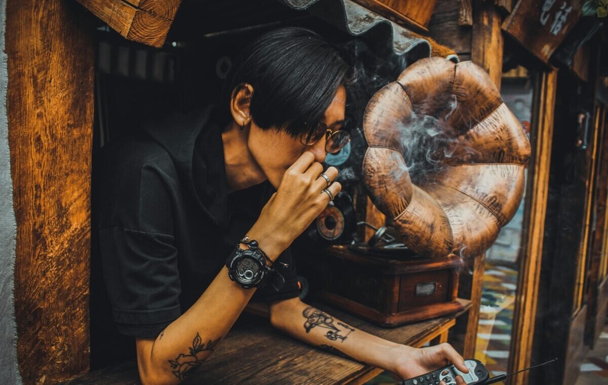 A person with tattoos is smoking while leaning out of a wooden window, with a vintage gramophone in the background, creating a relaxed and nostalgic atmosphere