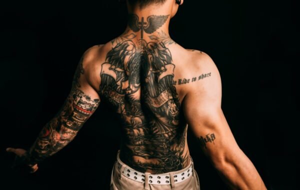 A person showing off a detailed tattoo design on their back and arms, featuring intricate artwork and phrases, posed against a dark background